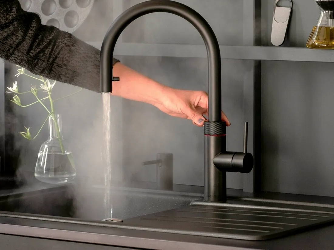 Quooker-Armatur in Schwarz: kochendes Wasser läuft dampfend in die Spüle, eine Hand bedient den Sicherheitsring